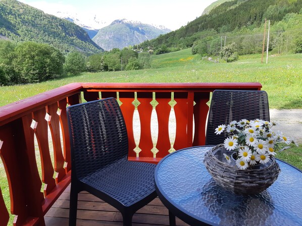 Una terraza con dos sillas y una mesa, una cesta de mimbre con margaritas, y montañas al fondo.