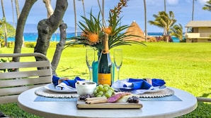 A table set with a bottle of wine, glasses, a plate with food, and a vase with flowers, set against a lush green lawn and palm trees, with a building and ocean in the background. - Napili Shores G157 Studio Bedroom Condo by RedAwning (Lahaina)