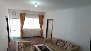 A living room with a sofa, a coffee table, and a patterned carpet. There is a window with curtains, a door, and a clock on the wall. - Hotel Salma (Bukittinggi)