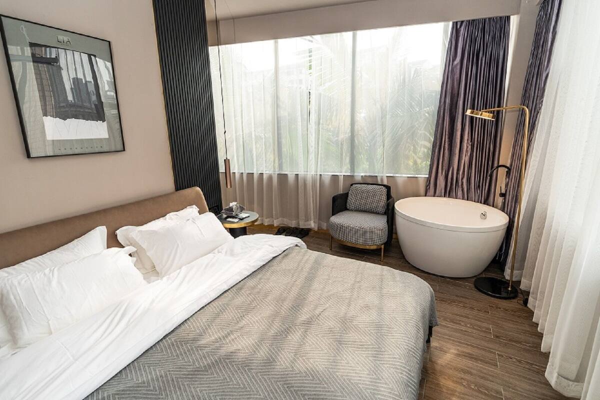 Habitación de hotel con una cama grande, una silla, una bañera y un ventanal con cortinas.
