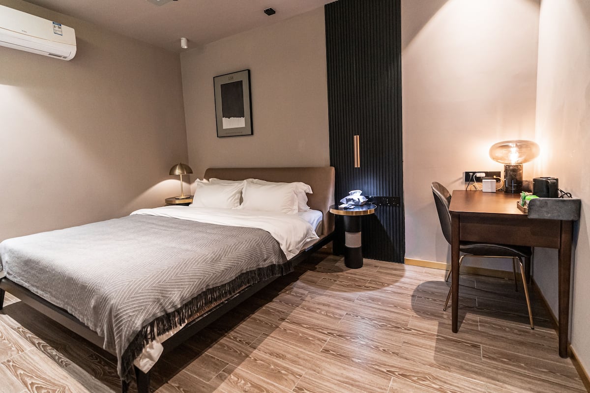 Habitación de hotel con cama, mesita de noche, lámpara, escritorio, silla y aire acondicionado de pared.