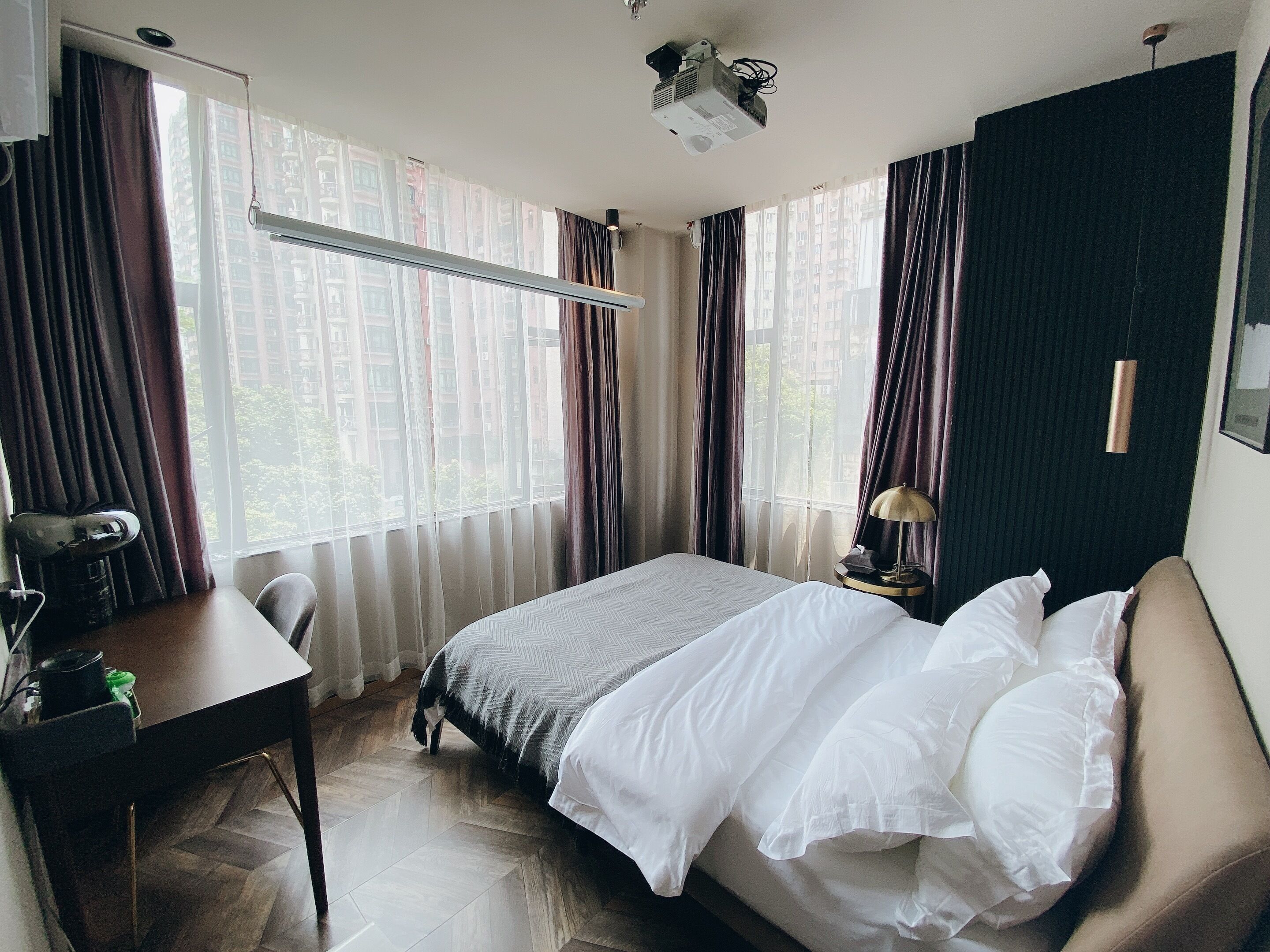 A hotel room with a bed, a desk, a chair, a lamp, and a projector on the ceiling.