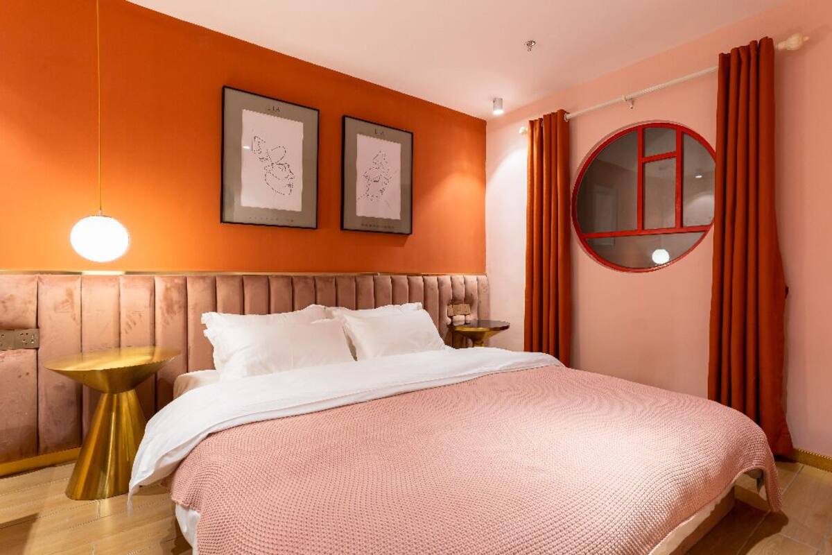 Un dormitorio con cama, dos cuadros enmarcados en la pared, un ventanal redondo con marco rojo y cortinas naranjas.