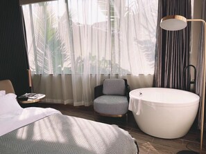 A hotel room with a large bed, a round bathtub, a small armchair, a bedside table, and sheer curtains allowing natural light. - LIA HOTEL (Shenzhen)