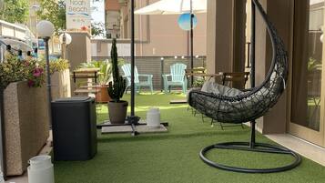 Un patio con césped artificial, una mecedora colgante y un sofá de mimbre blanco.