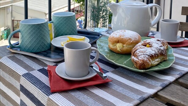 Un balcon avec une table dressée pour le petit-déjeuner, comprenant une théière, des tasses et des viennoiseries, donnant sur une plage et des bâtiments.