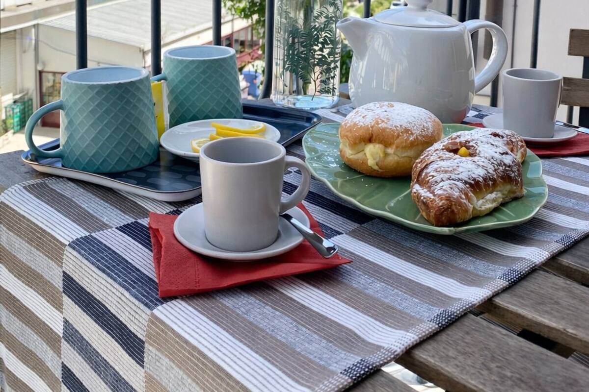 A balcony with a table set for breakfast, including a teapot, cups, and pastries, overlooking a beach and buildings.
