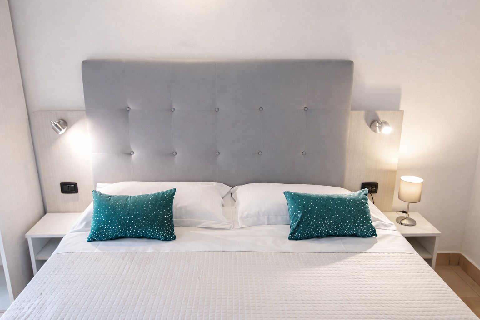 A neatly made bed with a tufted headboard, two white pillows, and two teal pillows with white speckles. There are two bedside tables with lamps on either side of the bed.