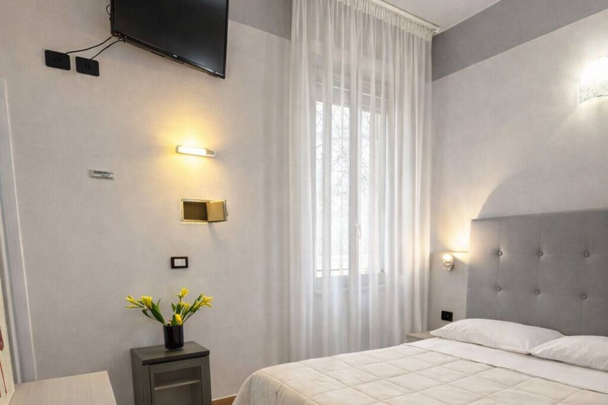 A bedroom with a bed, a TV mounted on the wall, a bedside table with a lamp, and a window with curtains.