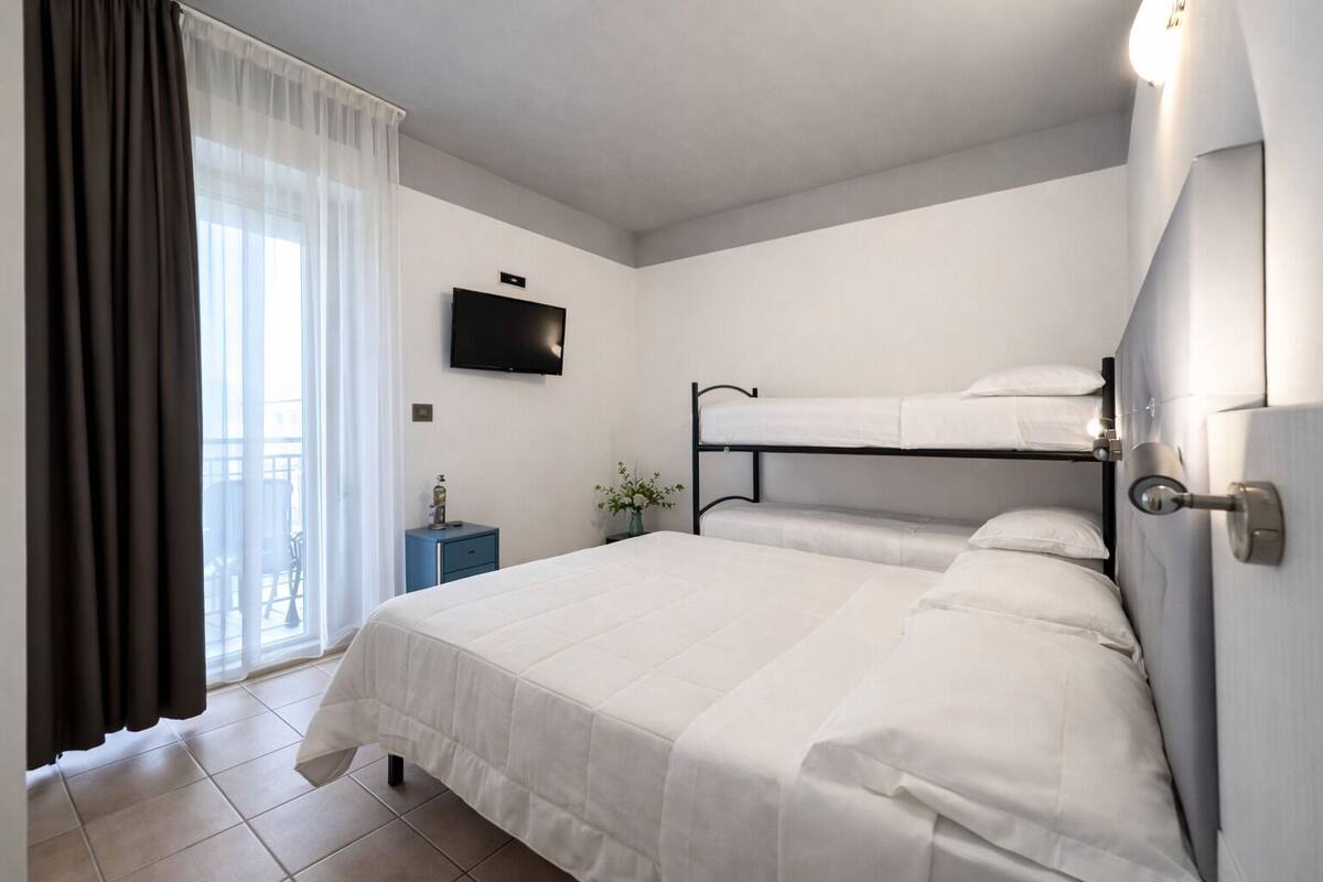 A hotel room with a bunk bed, a flat-screen TV, a bedside table with a lamp, and a view of the outdoors through a window with curtains.