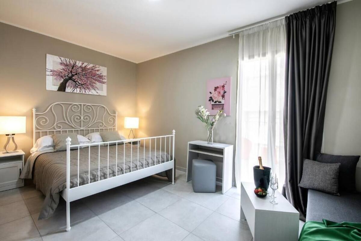 A bedroom with a bed, bedside tables, a nightstand, a dresser, a window with curtains, and wall art.