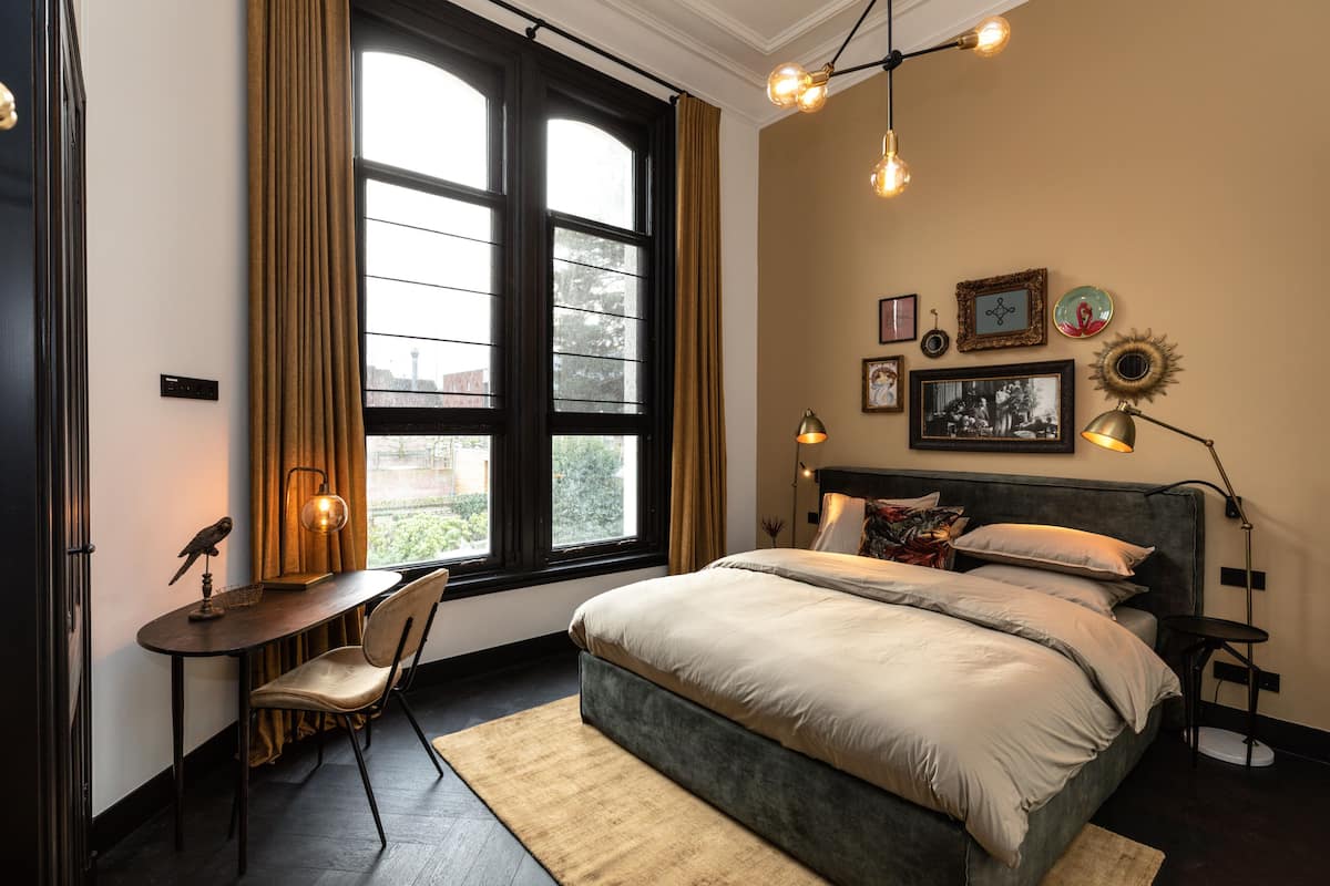 A bedroom with a bed, a desk, a chair, a window with curtains, and wall-mounted lights.