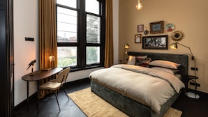 Una camera da letto con una grande finestra, una scrivania con una sedia, un letto con cuscini e lampade a parete.