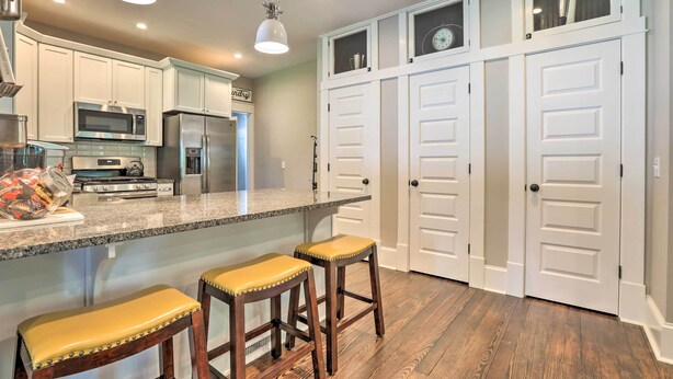 A modern kitchen with white cabinets, a granite countertop, and stainless steel appliances. There are two wooden stools with yellow cushions and a pendant light above the counter. - Renovated Craftsman House w/ Patio & Fire Pit! (Vevay)