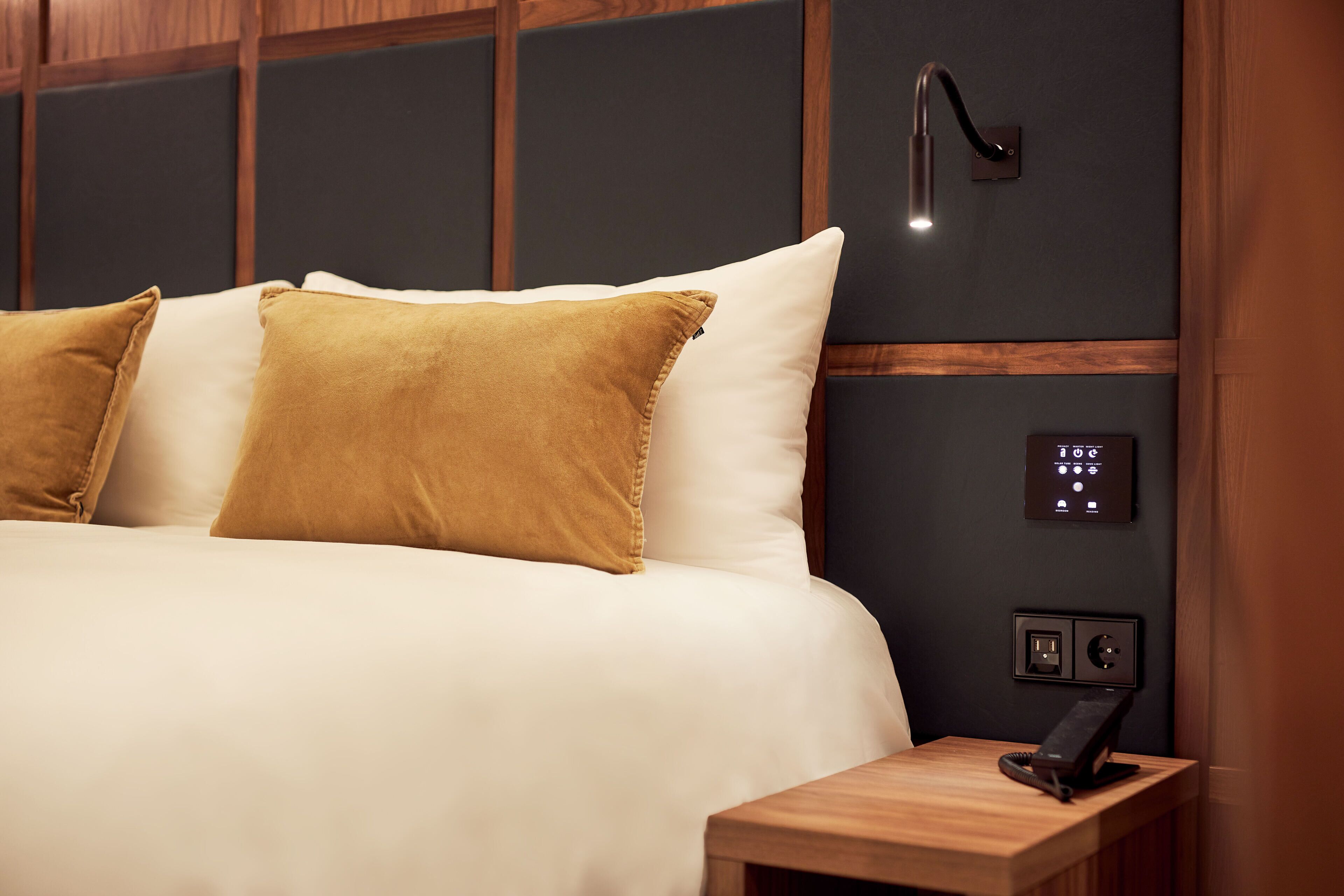 A neatly made bed with white and mustard pillows, a bedside table with a phone, and a wall-mounted control panel.