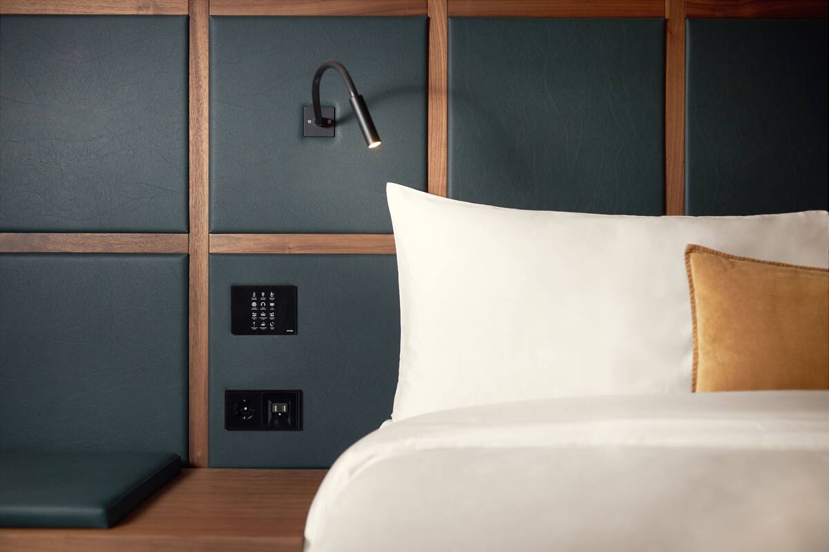 A hotel room with a bed, a bedside table, a wall-mounted light, and a control panel.