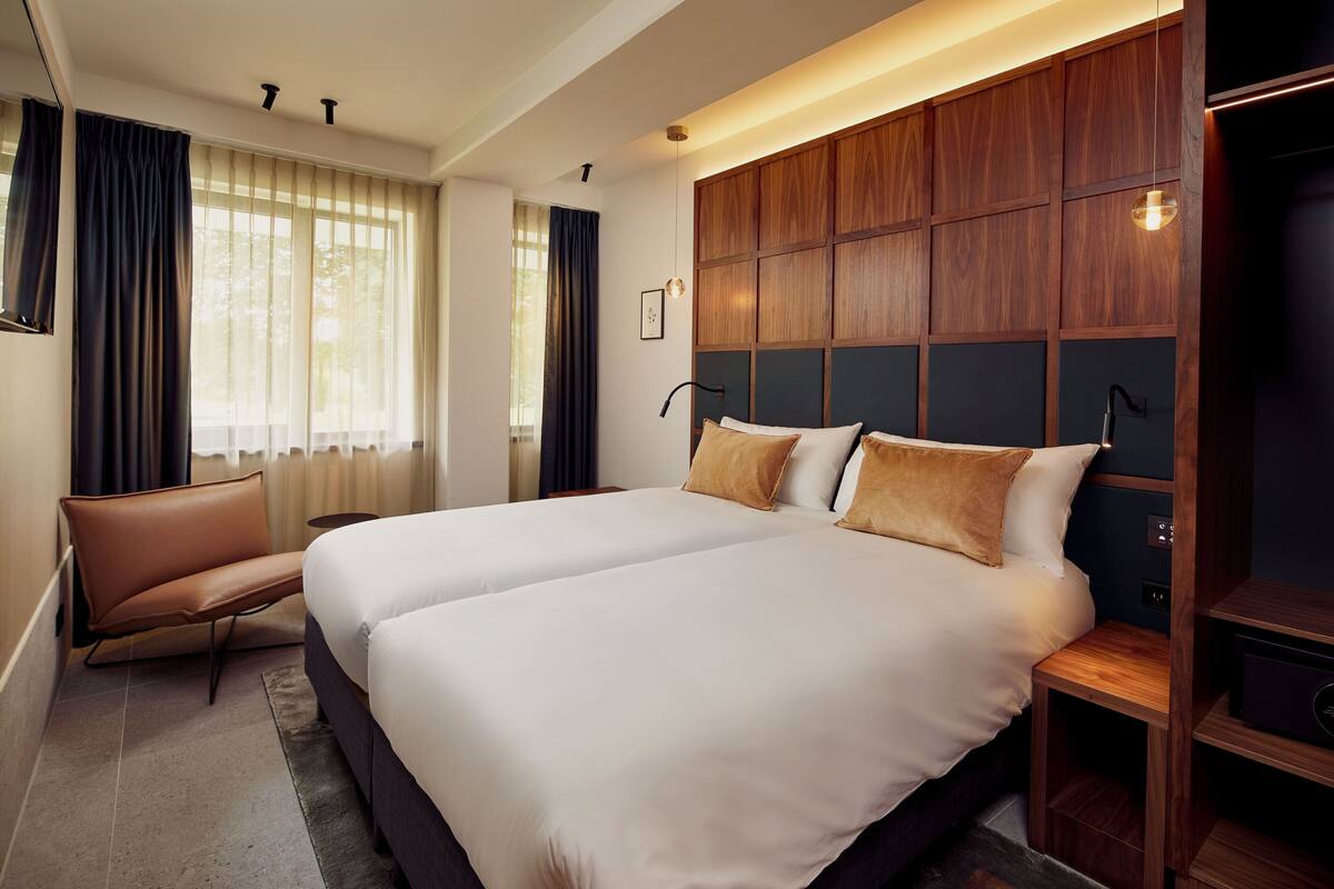 A modern hotel room with two beds, a leather chair, wooden paneling, and a nightstand.