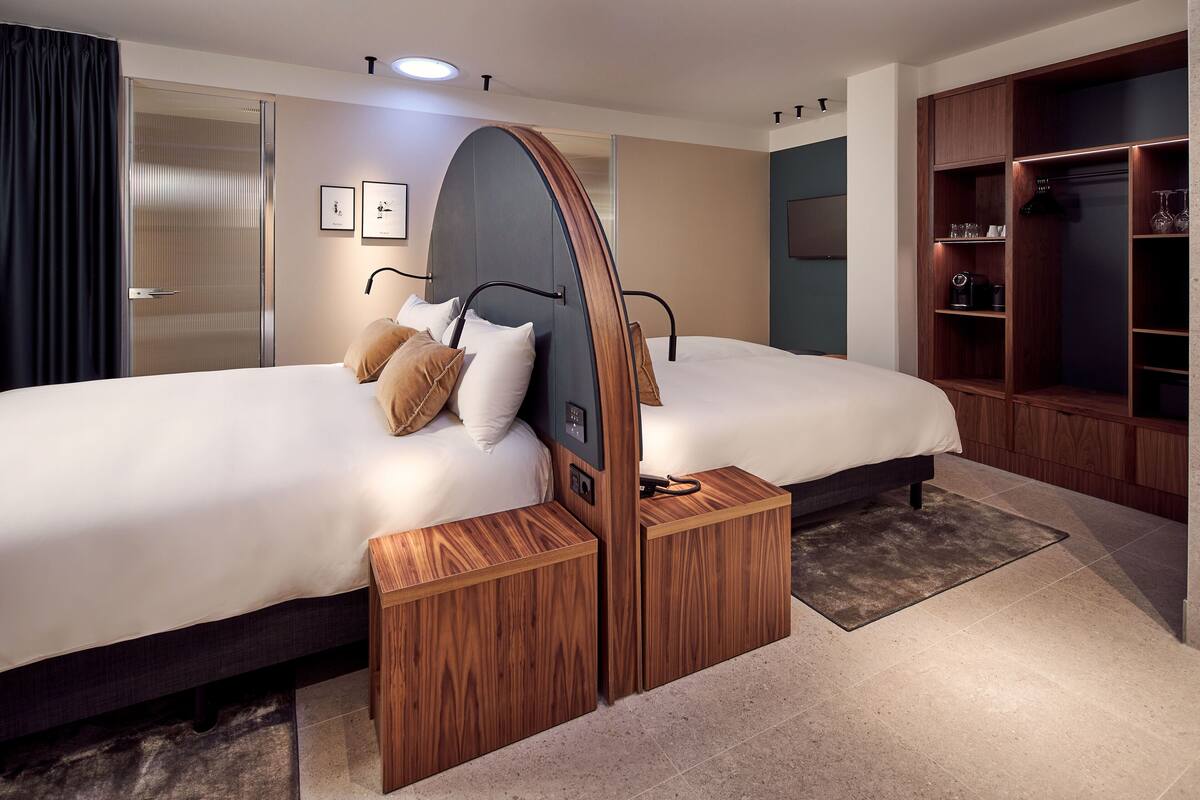 A modern hotel room with two beds, a wooden wardrobe, and a small seating area.