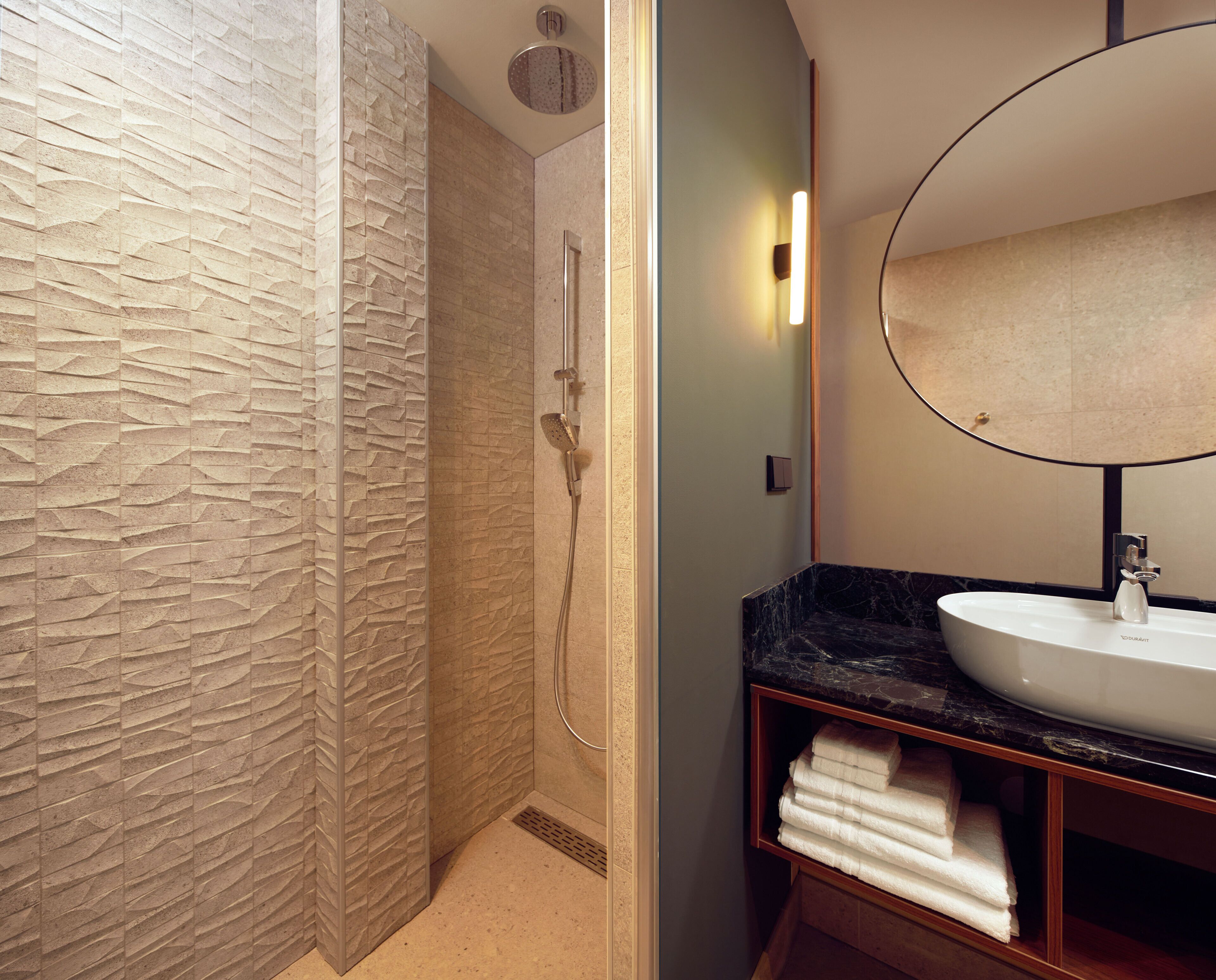 A bathroom with a stone-textured shower wall, a white sink with a marble countertop, and a wooden vanity with towels.