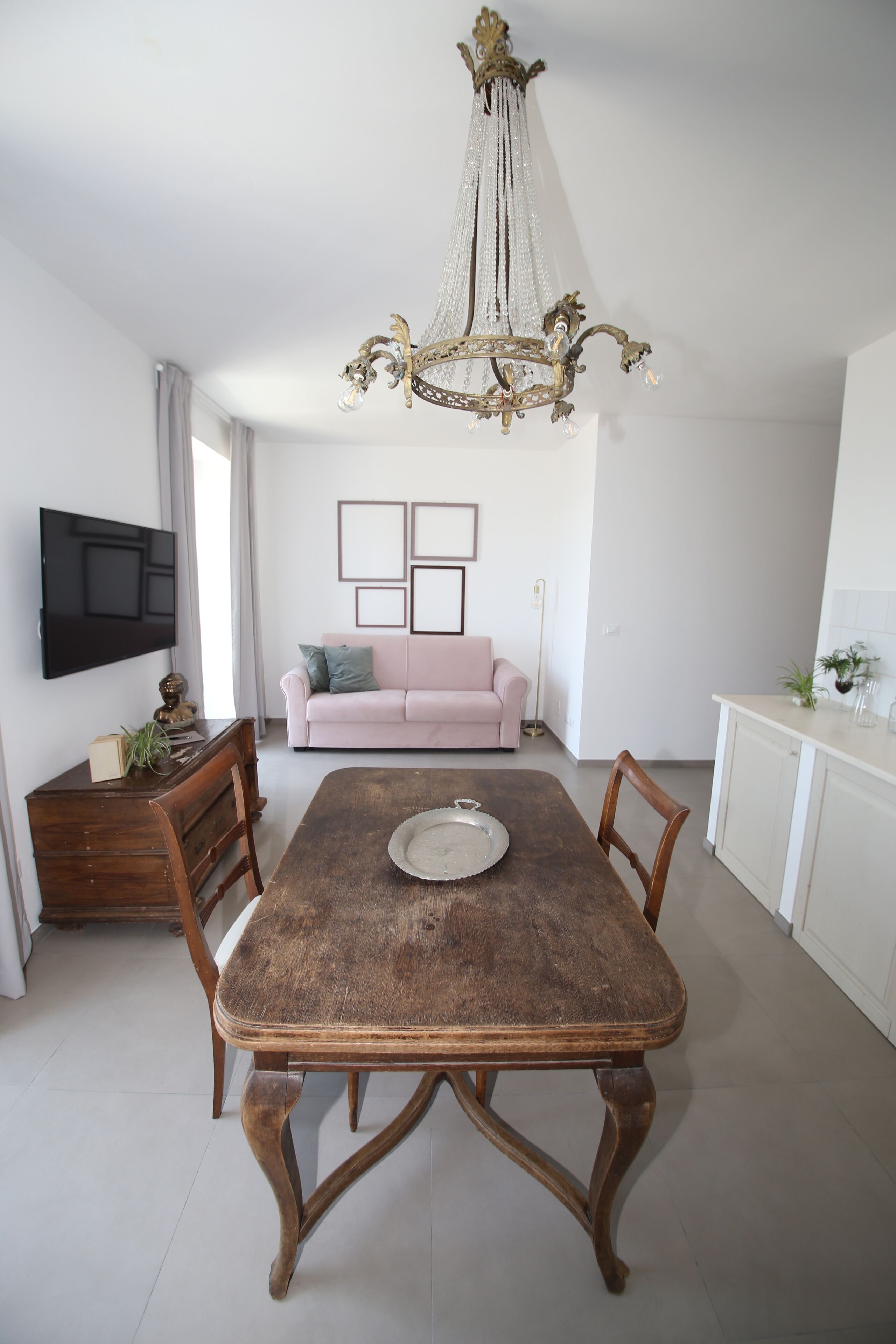 A dining area with a wooden table, chairs, a chandelier, a pink sofa, and a TV on the wall.