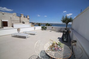 A rooftop terrace with a table set for dining, lounge chairs, and a view of the sea. - Don Alfredo (Taranto)