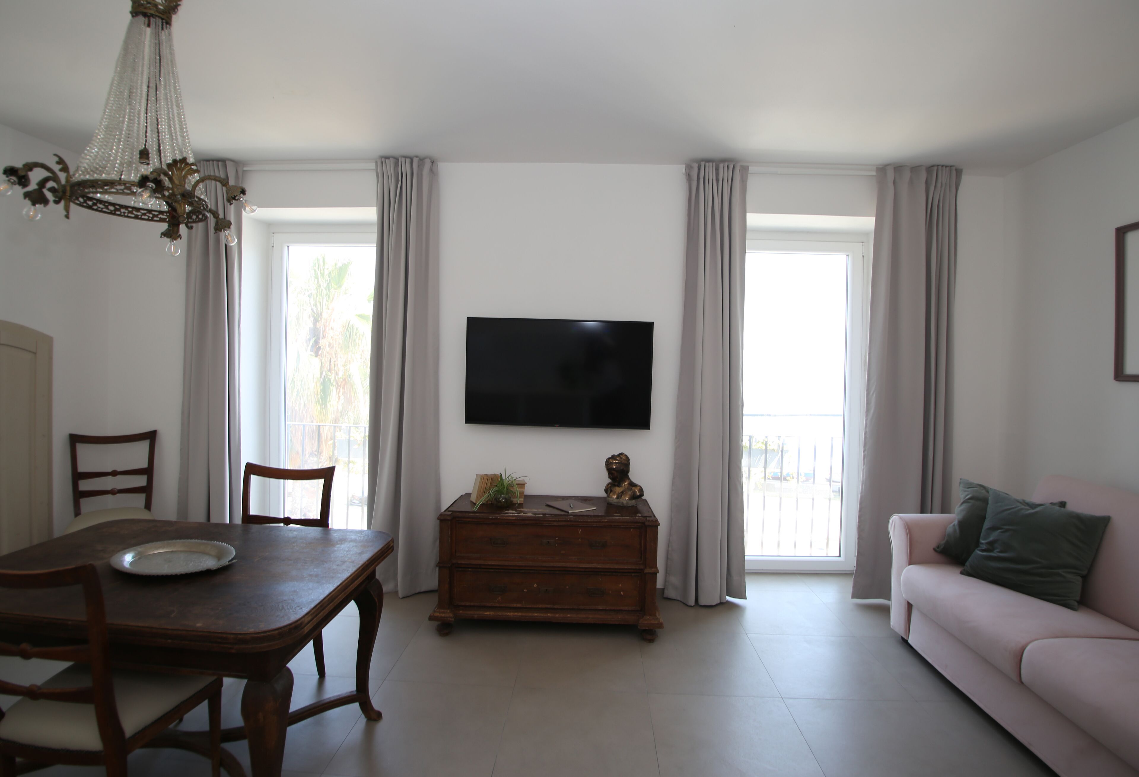 A living room with a dining table, chairs, a sofa, a TV mounted on the wall, and a chandelier.