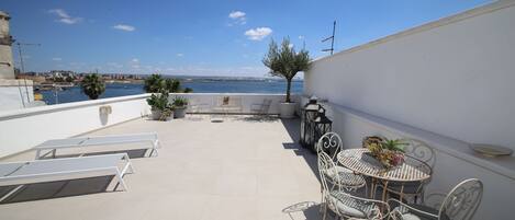 A rooftop terrace with a table and chairs, potted plants, and a view of the sea.