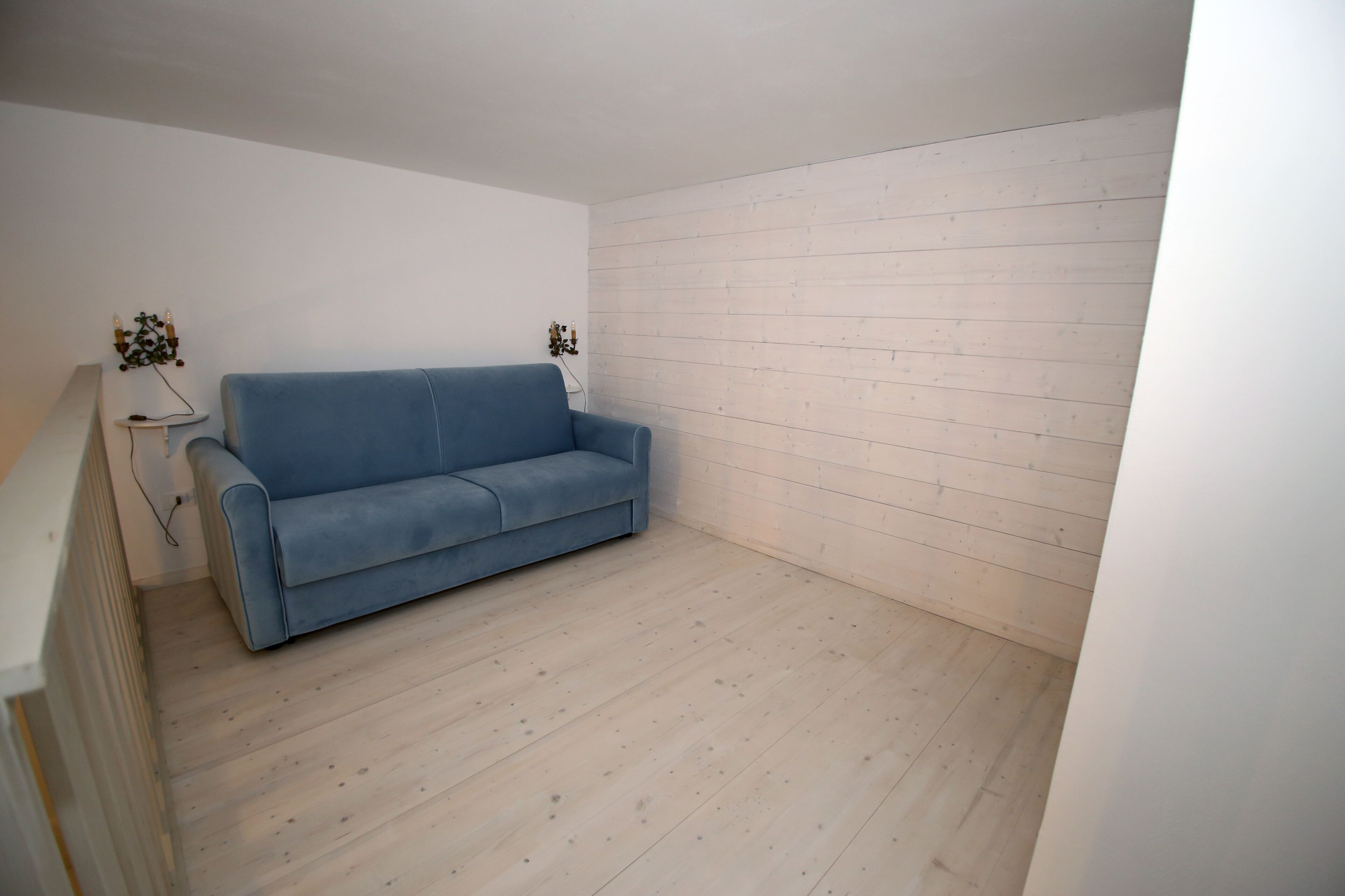 Una habitación con un sofá azul frente a una pared blanca, piso de madera y una mesita con una lámpara.