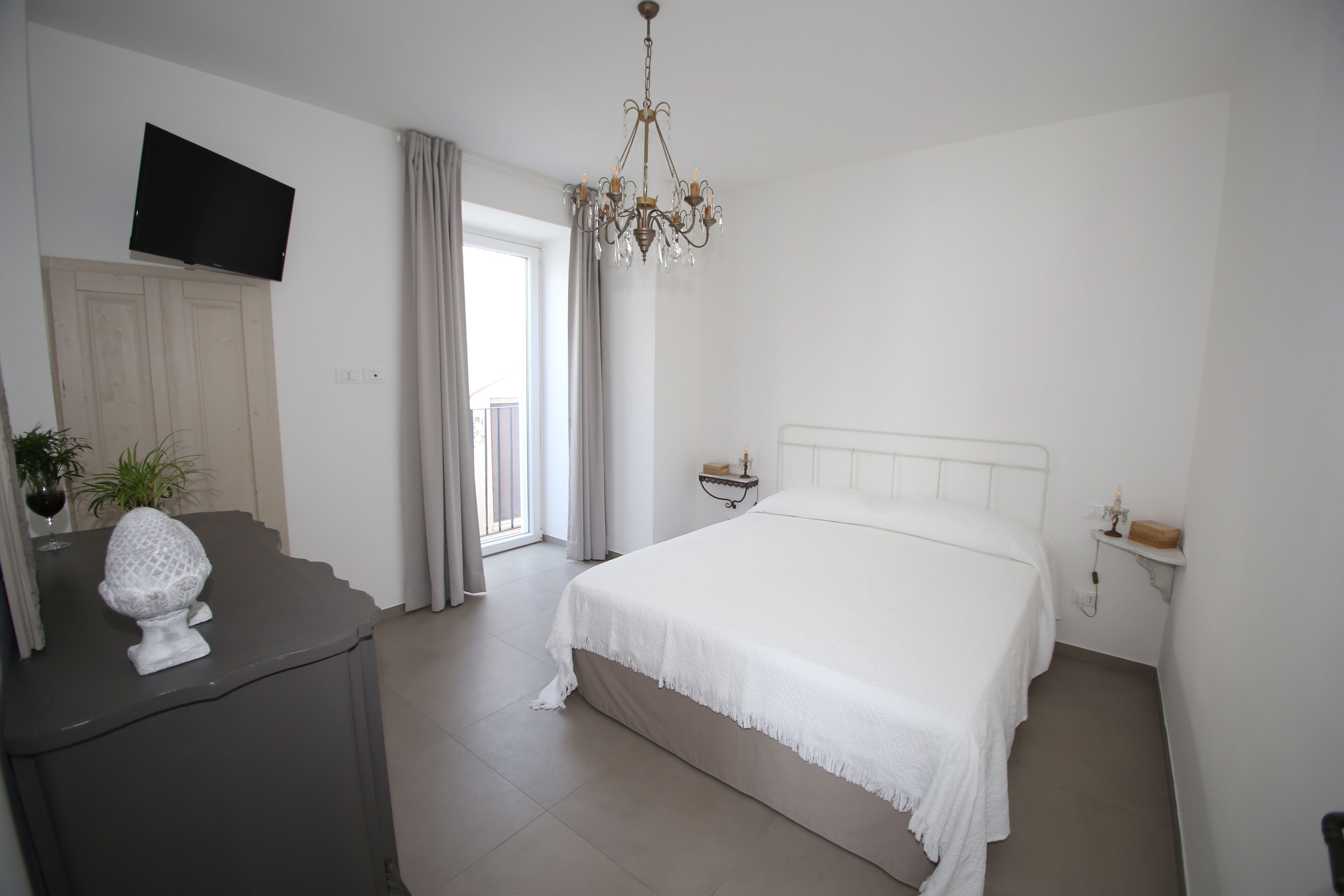 Une chambre à coucher comprenant un lit, un téléviseur fixé au mur, un lustre, une table de chevet et une fenêtre avec des rideaux.