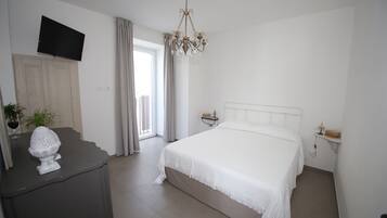 Un dormitorio con cama, televisor montado en la pared, candelabro, mesita de noche y ventana con cortinas.