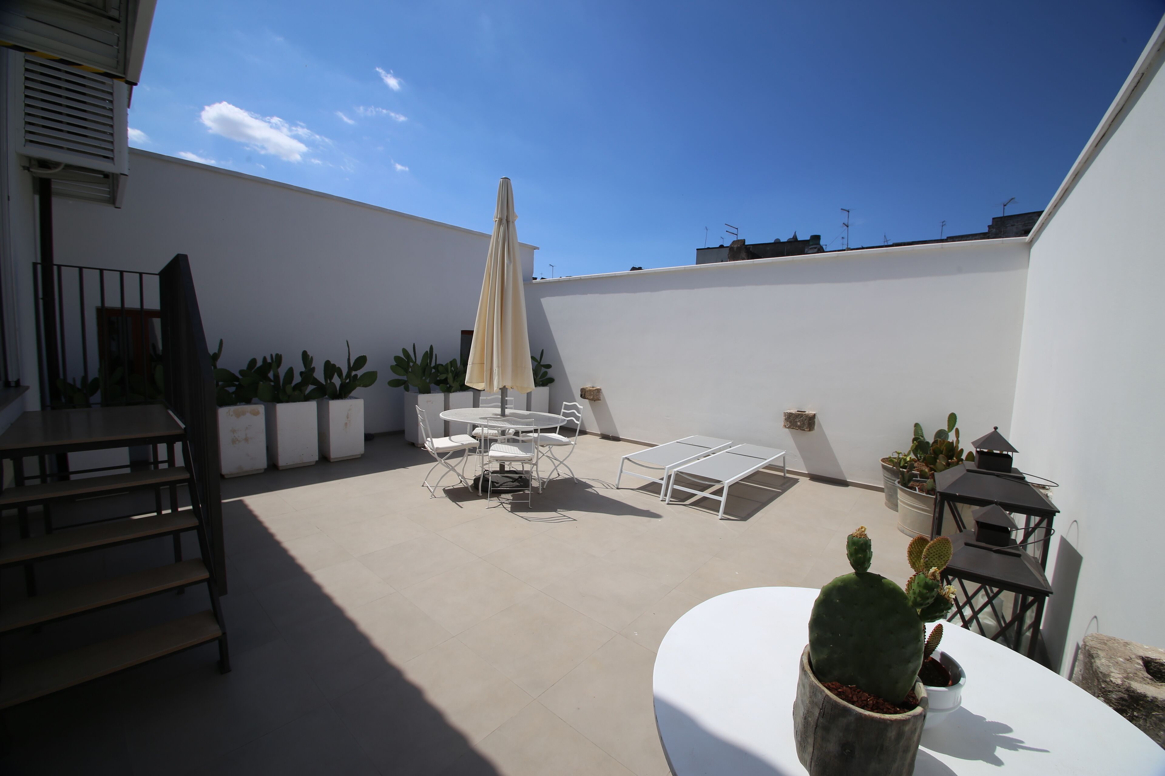 Un patio al aire libre con una mesa y sillas blancas, plantas en macetas y un paraguas grande.