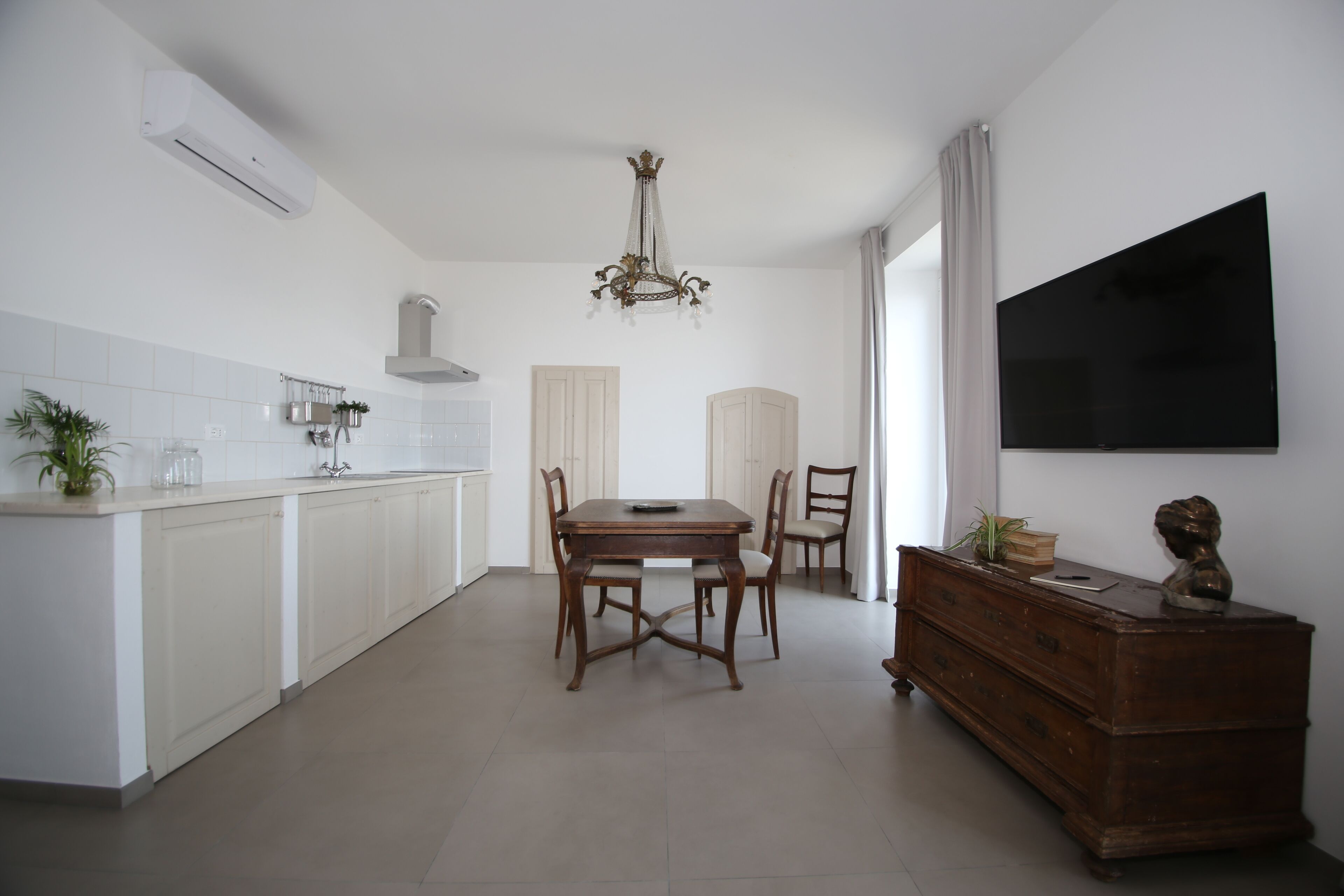 A modern kitchen with white cabinetry, a dining table with chairs, a chandelier, and a flat-screen TV mounted on the wall.