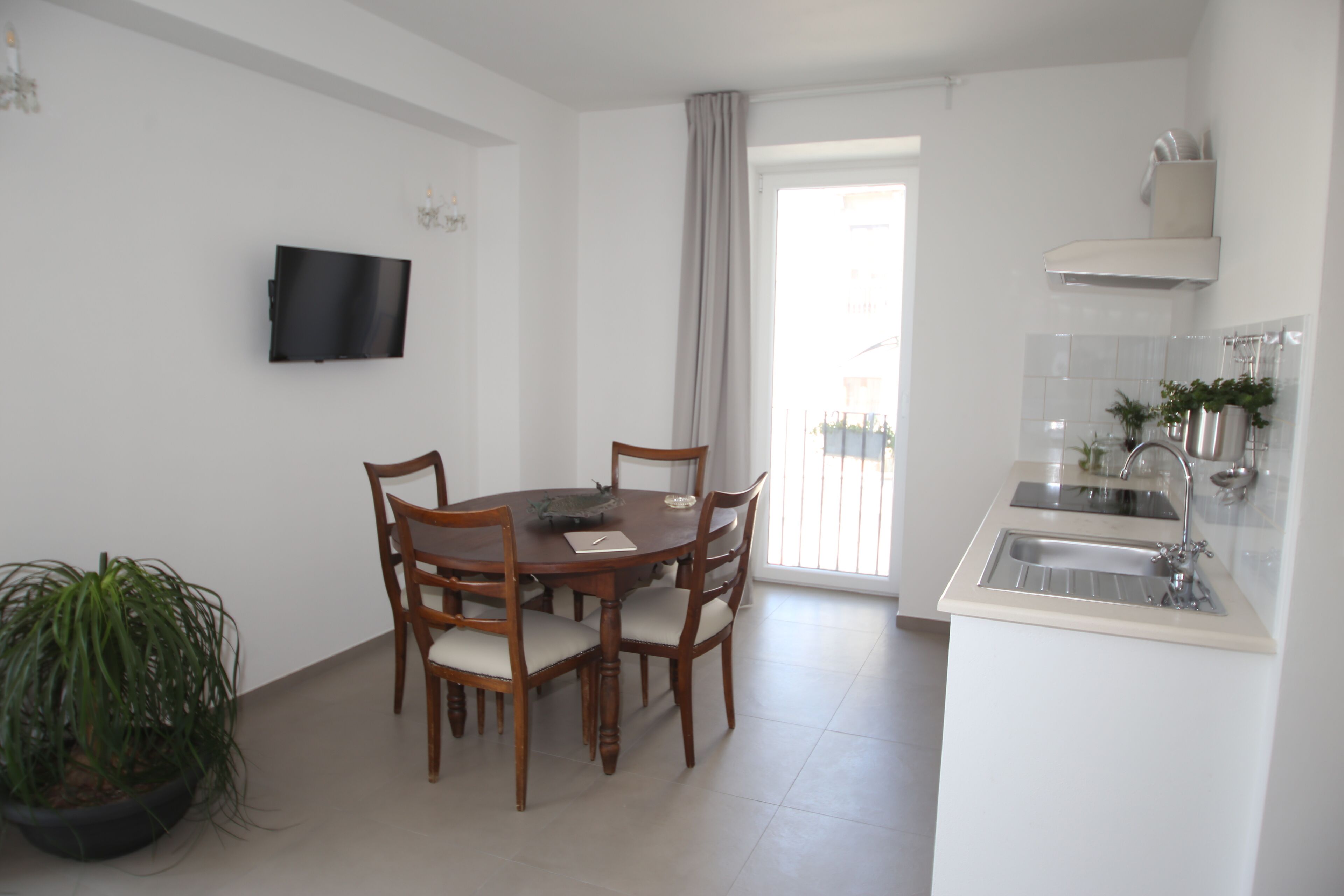 Un comedor con una mesa redonda y sillas, una planta en maceta, un televisor de pantalla plana en la pared y una cocina con un lavaplatos y armarios.