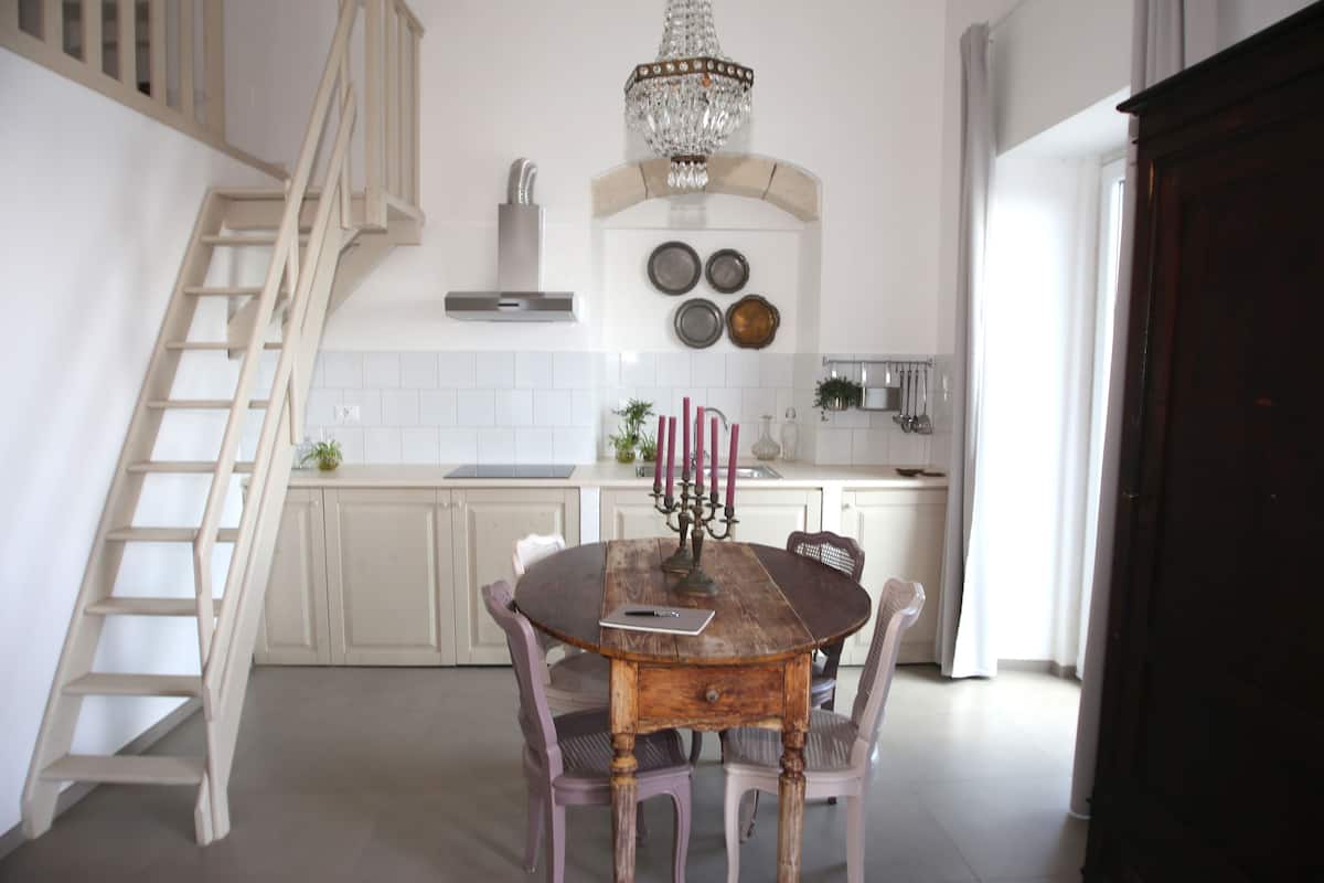 A dining area with a wooden table, four chairs, a chandelier, and a kitchen in the background.