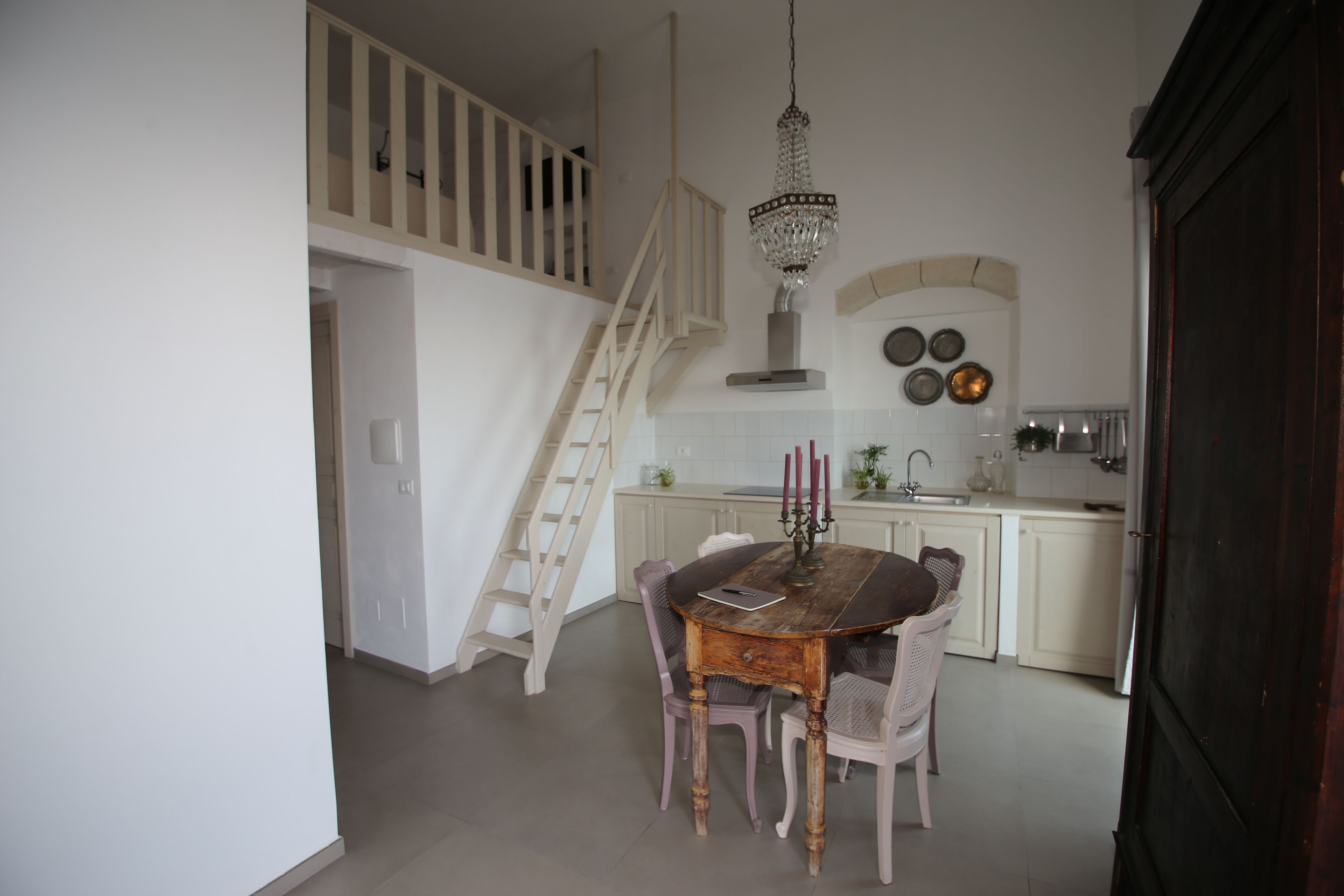A dining area with a wooden table and chairs, a chandelier, a staircase, and a kitchen in the background.