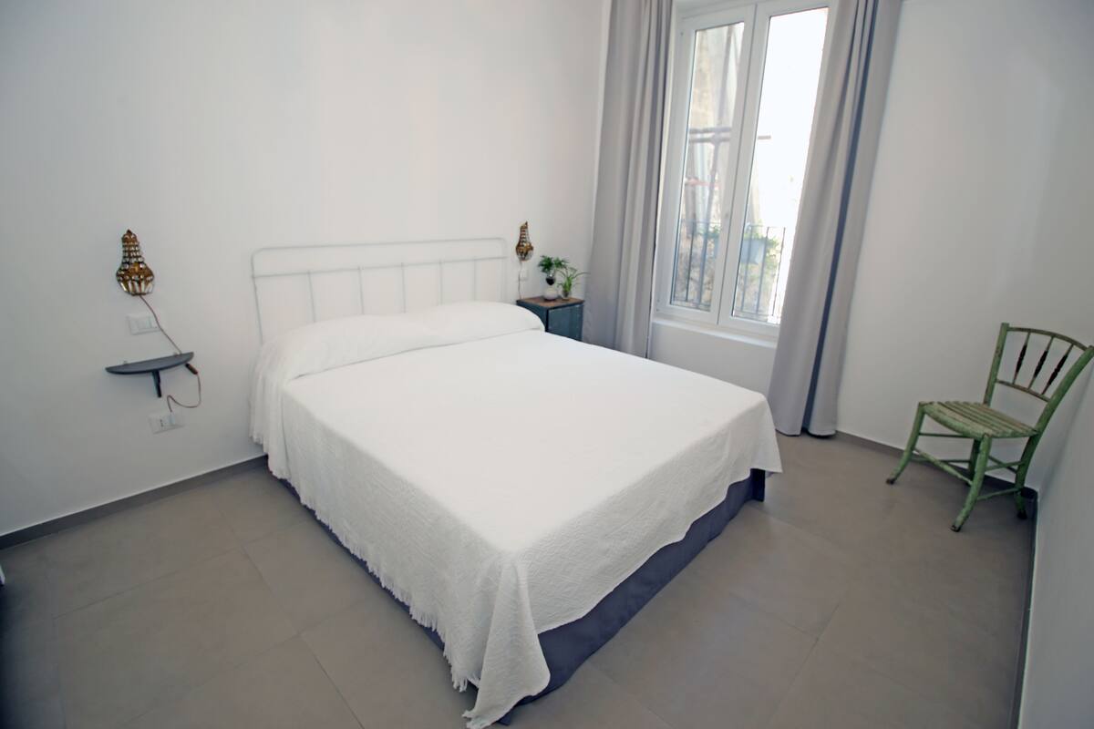 A neatly made bed with a white bedspread in a room with a window, a small bedside table with a lamp, and a green chair.