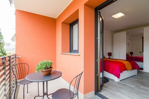 A balcony with a table and chairs, a potted plant, and a sliding glass door leading to a bedroom with a bed and a nightstand.