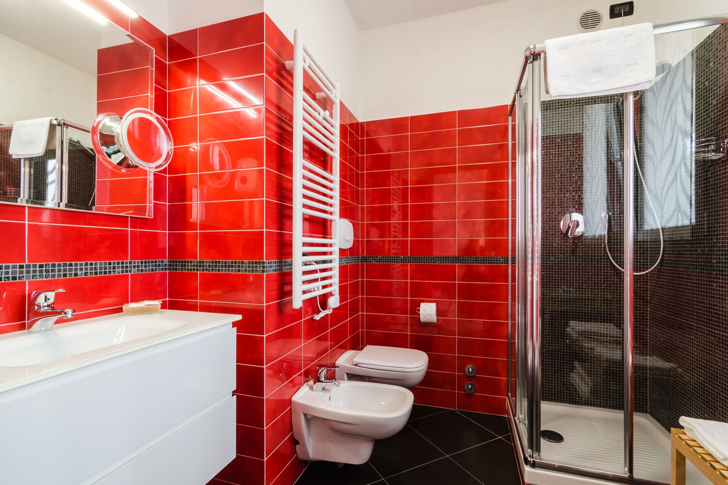 Ein Badezimmer mit roten Fliesen, einer weißen Badewanne, zwei Toiletten, einem Spiegel und einer Duschkabine aus Glas.