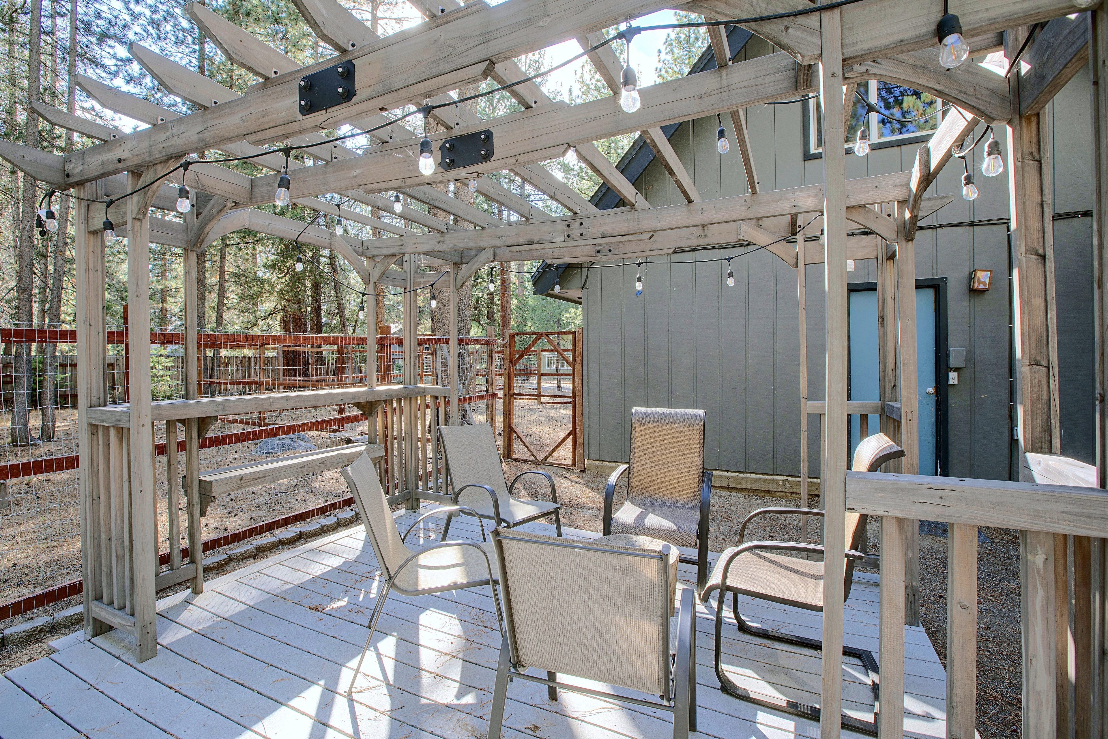 Une terrasse en bois, dotée de chaises et d’une structure en forme de pergola, est entourée d’une zone délimitée par une clôture, avec des arbres en arrière-plan.