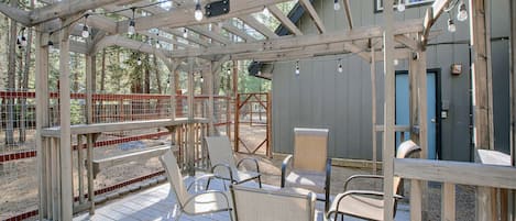 A wooden deck with chairs and a pergola structure, surrounded by a fenced area with trees in the background.