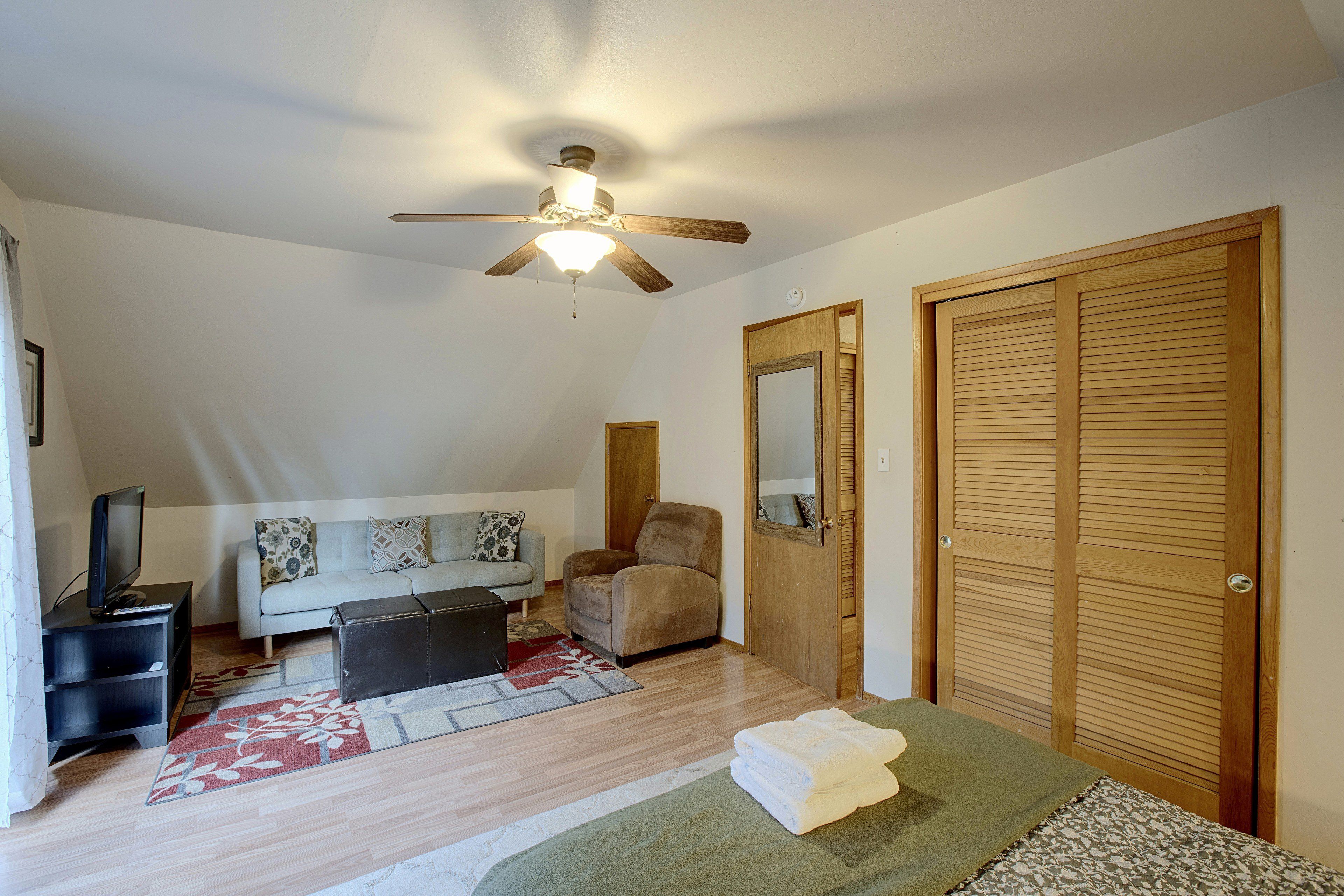 Une pièce avec un ventilateur de plafond, un canapé, un fauteuil, un téléviseur posé sur un meuble et une porte en bois avec un miroir.