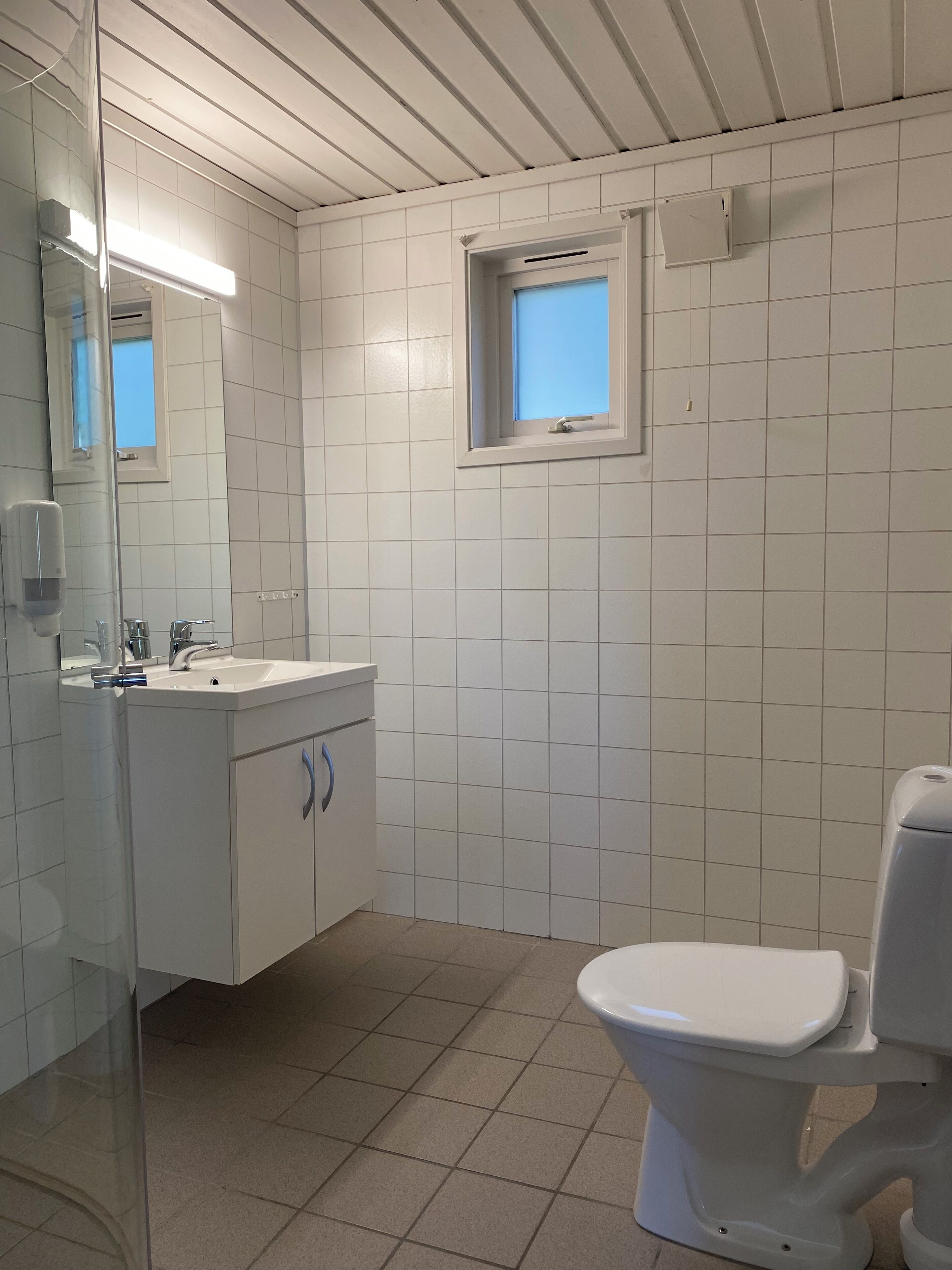 Un baño con azulejos blancos, un lavamanos con espejo, una ventana y un inodoro.