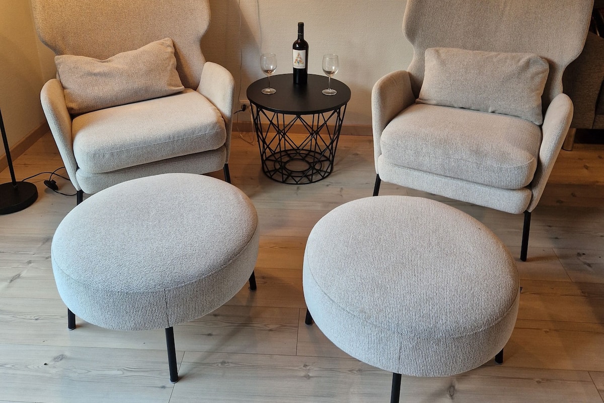 A cozy room with two armchairs and ottomans, a small table with a bottle and glasses, and a lamp.