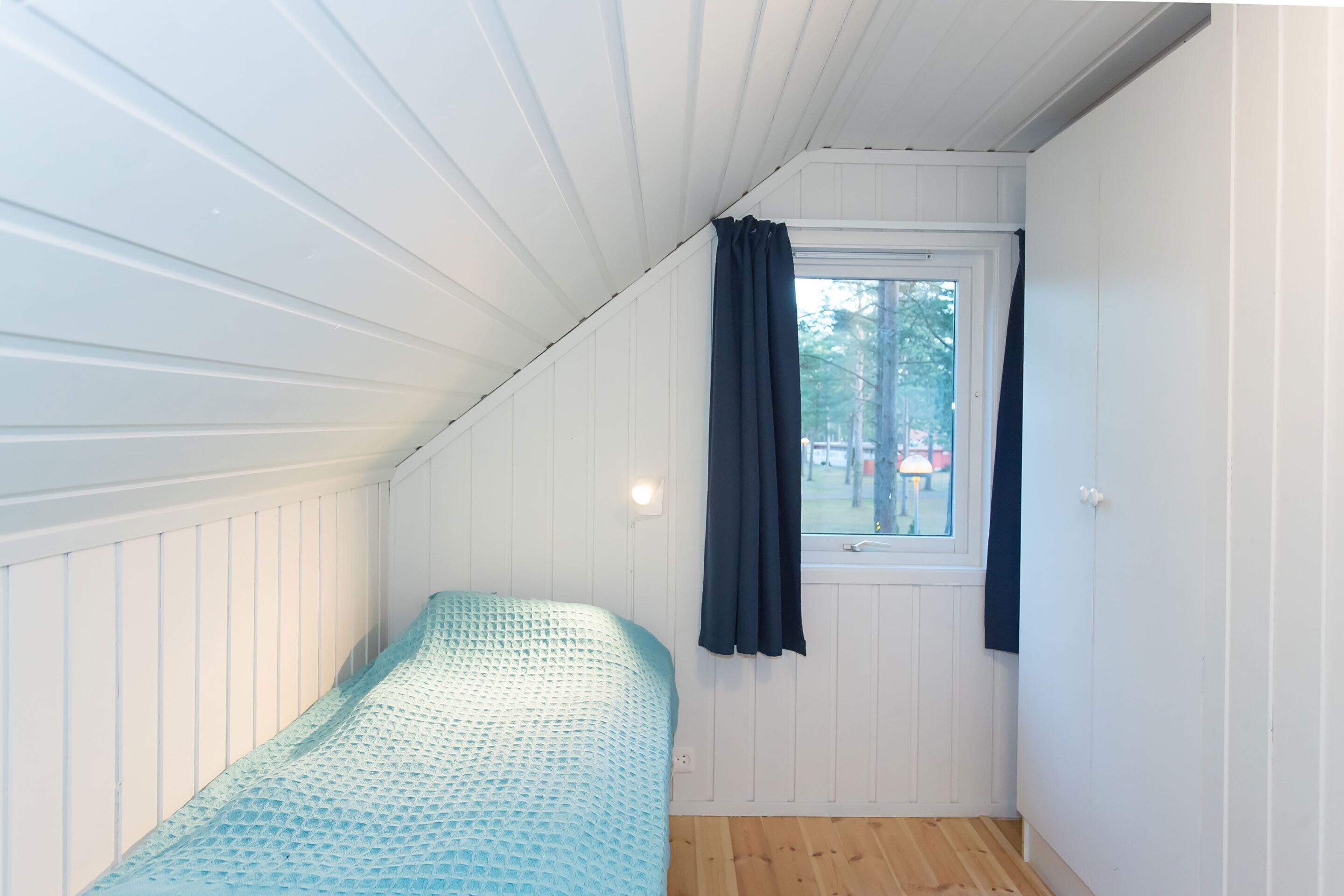 Una habitación pequeña con una cama individual, paredes blancas con paneles y techo inclinado.