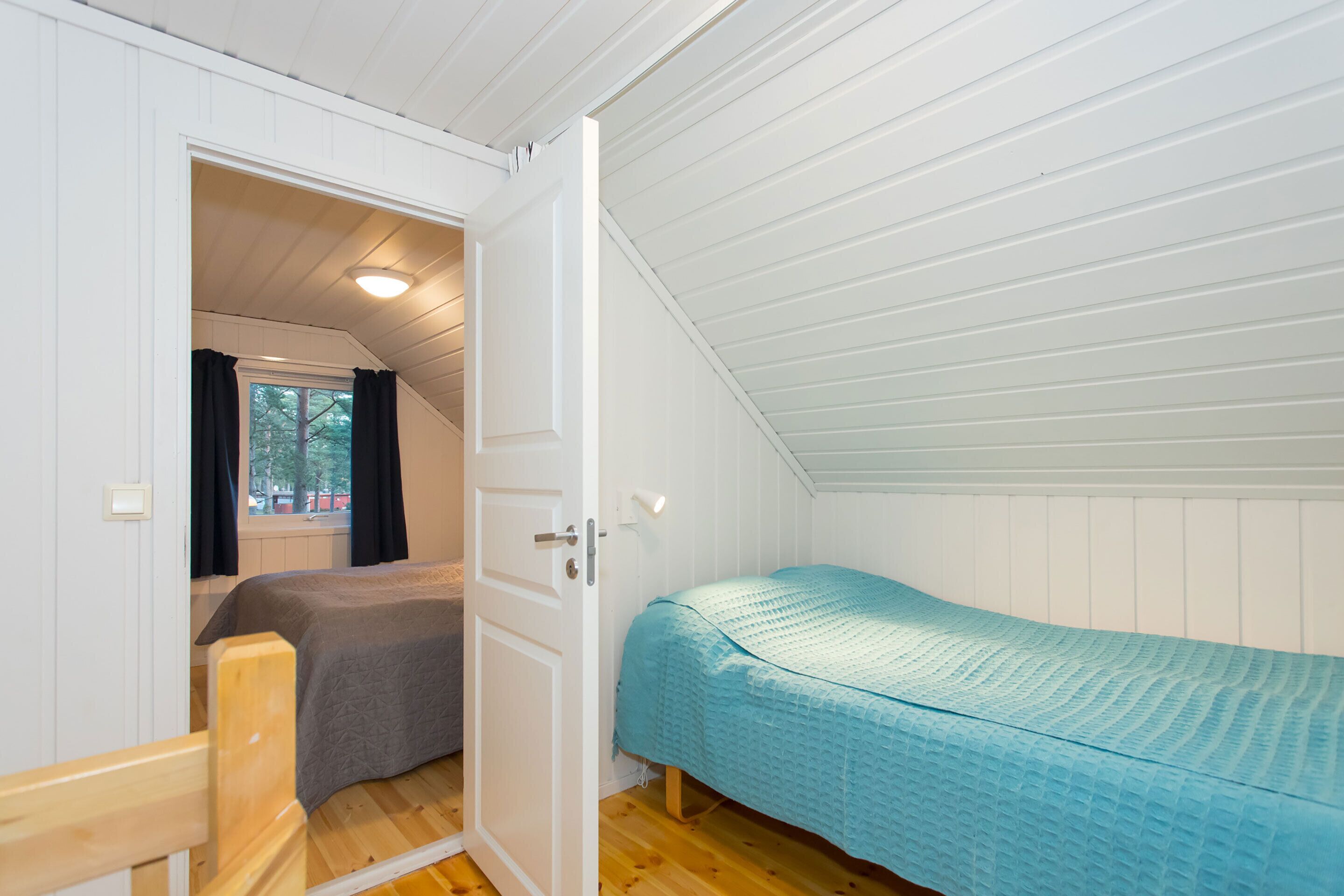 Eine kleine, einfache Unterkunft mit schrägem Dach, einem Bett mit blauer Bettdecke, einem Holzstuhl und einem Fenster mit schwarzen Vorhängen.