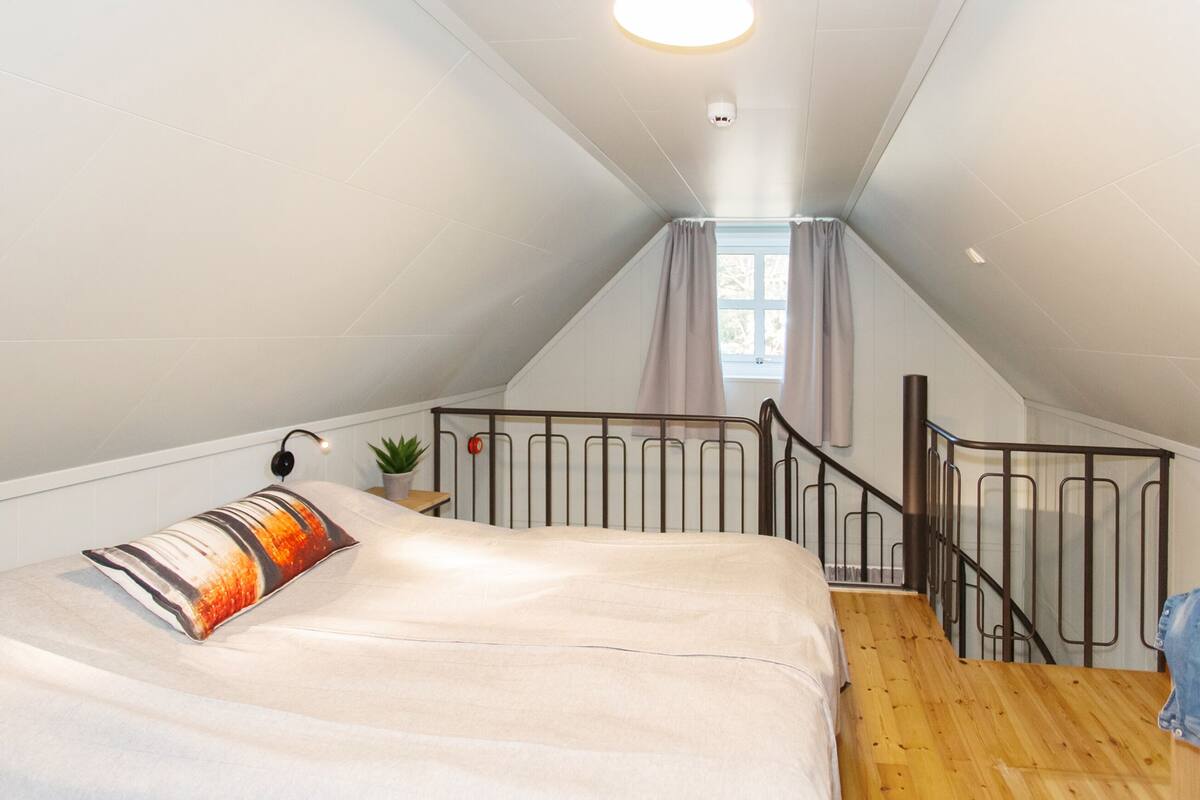 A bedroom with a bed, a small potted plant, a nightstand, a window with curtains, and a staircase leading to an upper level.