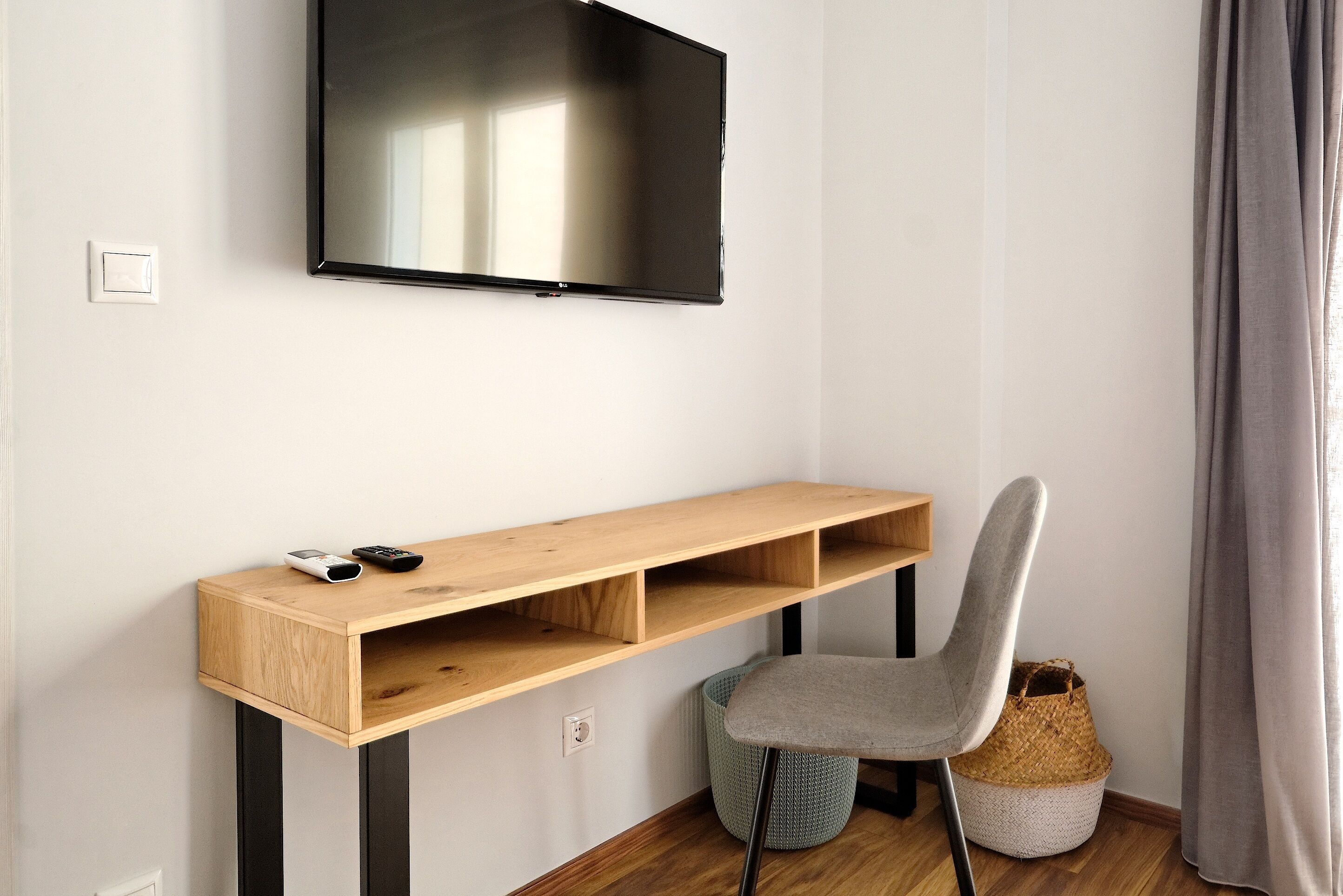 Une chambre moderne dotée d’un téléviseur à écran plat fixé au mur, d’un bureau en bois avec une chaise et d’un petit panier posé sur le sol.