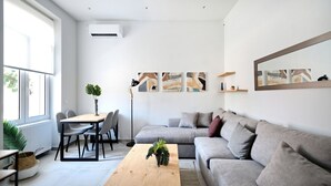 A modern living room with a grey sofa, a wooden coffee table, and a dining area with chairs and a table. There are abstract paintings on the wall and a large mirror above the sofa.