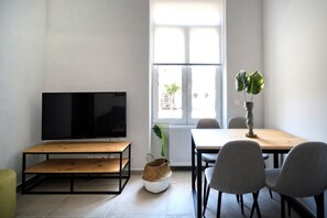 A modern living room with a flat-screen TV, a wooden table, two grey chairs, a small woven basket, and a potted plant.