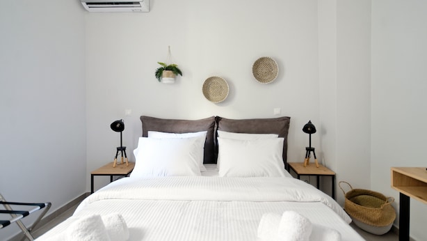 A neatly made bed with white linens, two bedside tables with lamps, a wall-mounted air conditioner, and decorative woven baskets.